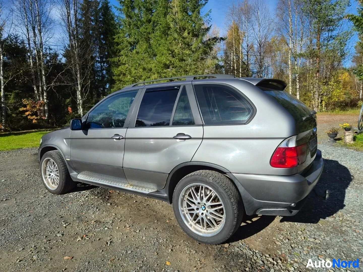 Takaviisto: Hopeanharmaa maastoauto pysäköitynä sorapintaiselle alueelle metsätaustan edessä, nähtynä takavasemmalta. Autossa on kattokiskot, takaspoileri ja vetokoukku.