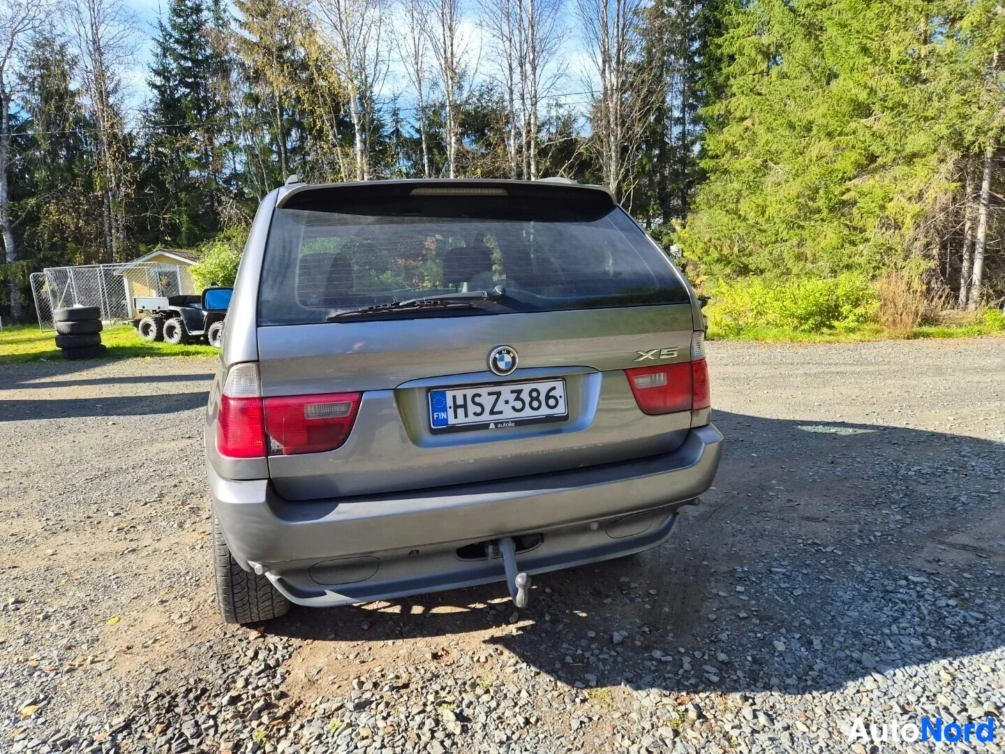 Takaa: Hopeanharmaa maastoauto pysäköitynä sorapintaiselle alueelle metsätaustan edessä, nähtynä takaa. Autossa on vetokoukku ja rekisterikilpi