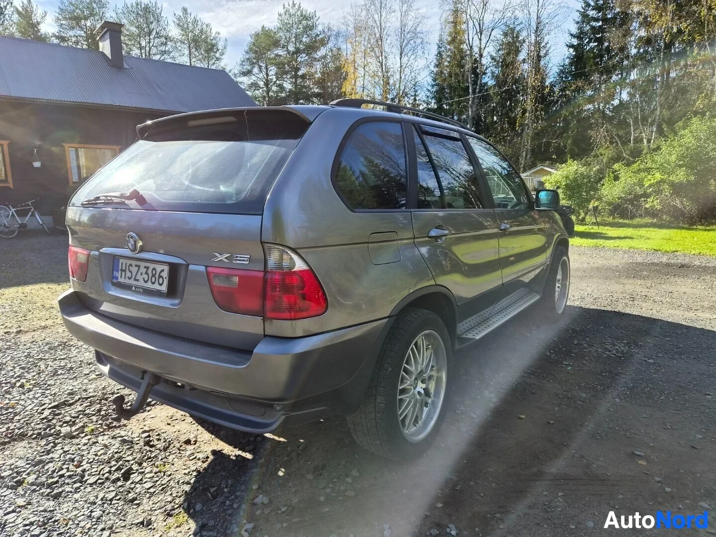 Takaviisto: Harmaa SUV pysäköitynä sepelipihalla. Ajoneuvon vasen takakulma on kuvassa, rekisterikilvessä teksti. Autossa on vetokoukku, kattokaiteet ja astinlaudat