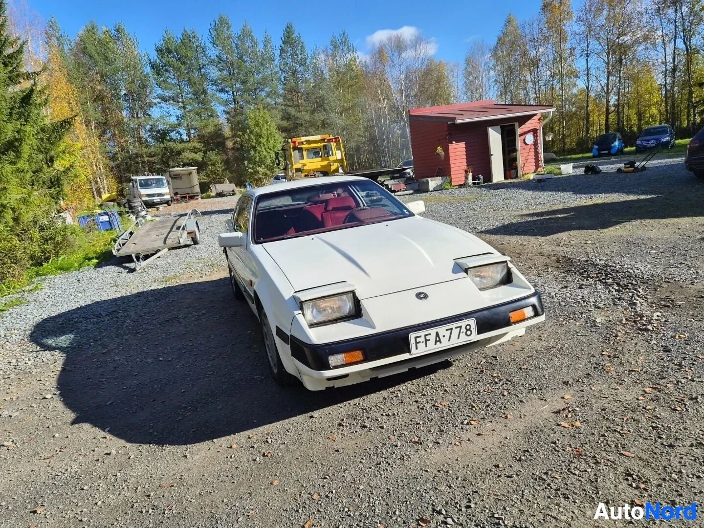 Etuviisto, nokka oikealle: Valkoinen kaksiovinen coupé, jossa on pop-up-ajovalot, pysäköitynä sorapihalla. Rekisterikilpi on näkyvissä ja punaiset istuimet näkyvät osittain