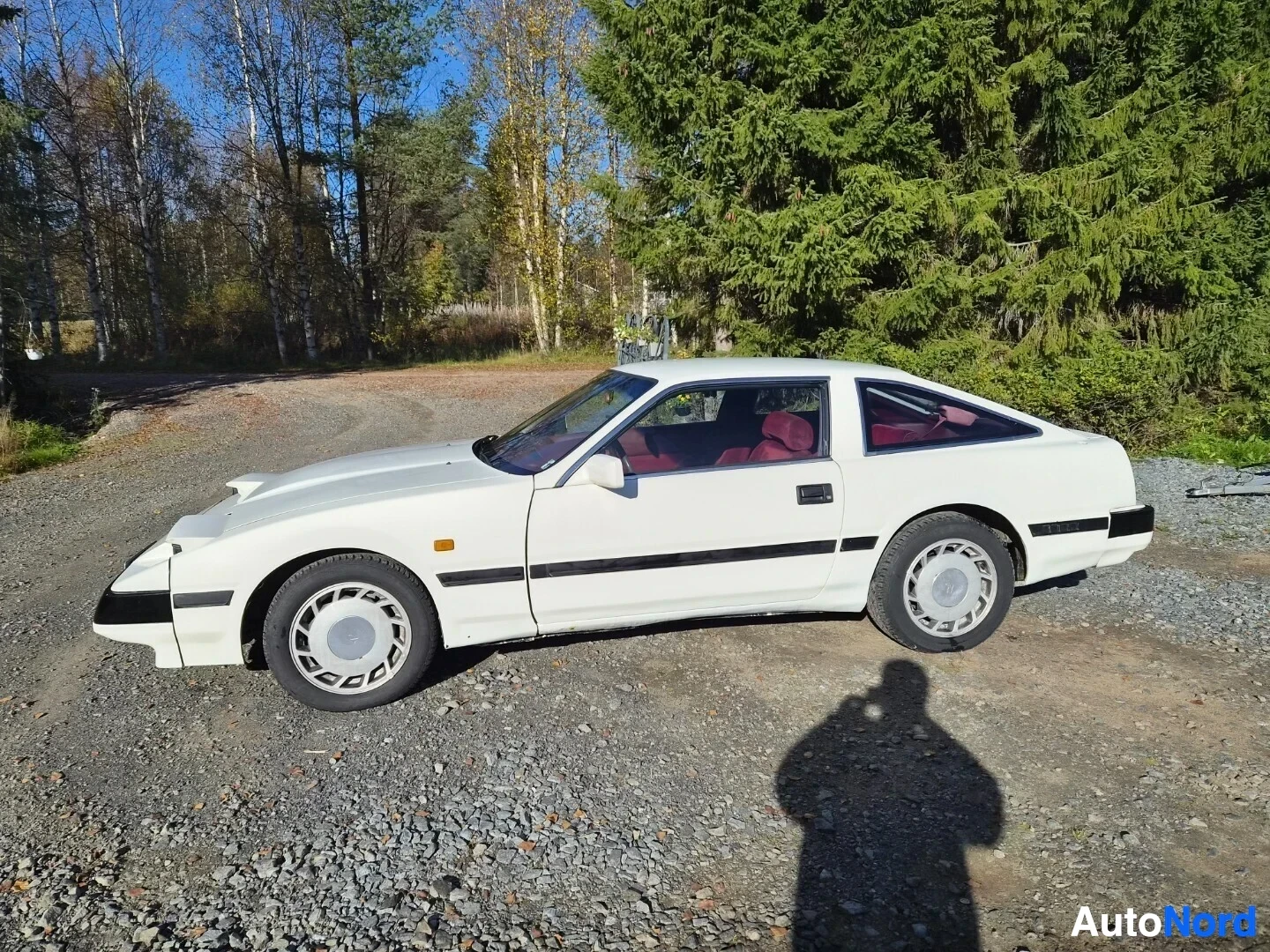 Sivulta: Valkoinen Nissan 300ZX kuvattu suoraan vasemmalta sivulta sorapihalla. Ajovalot ovat ylös nostetussa asennossa. Auto on sorapihalla, taustalla puita. Kuvaajan v