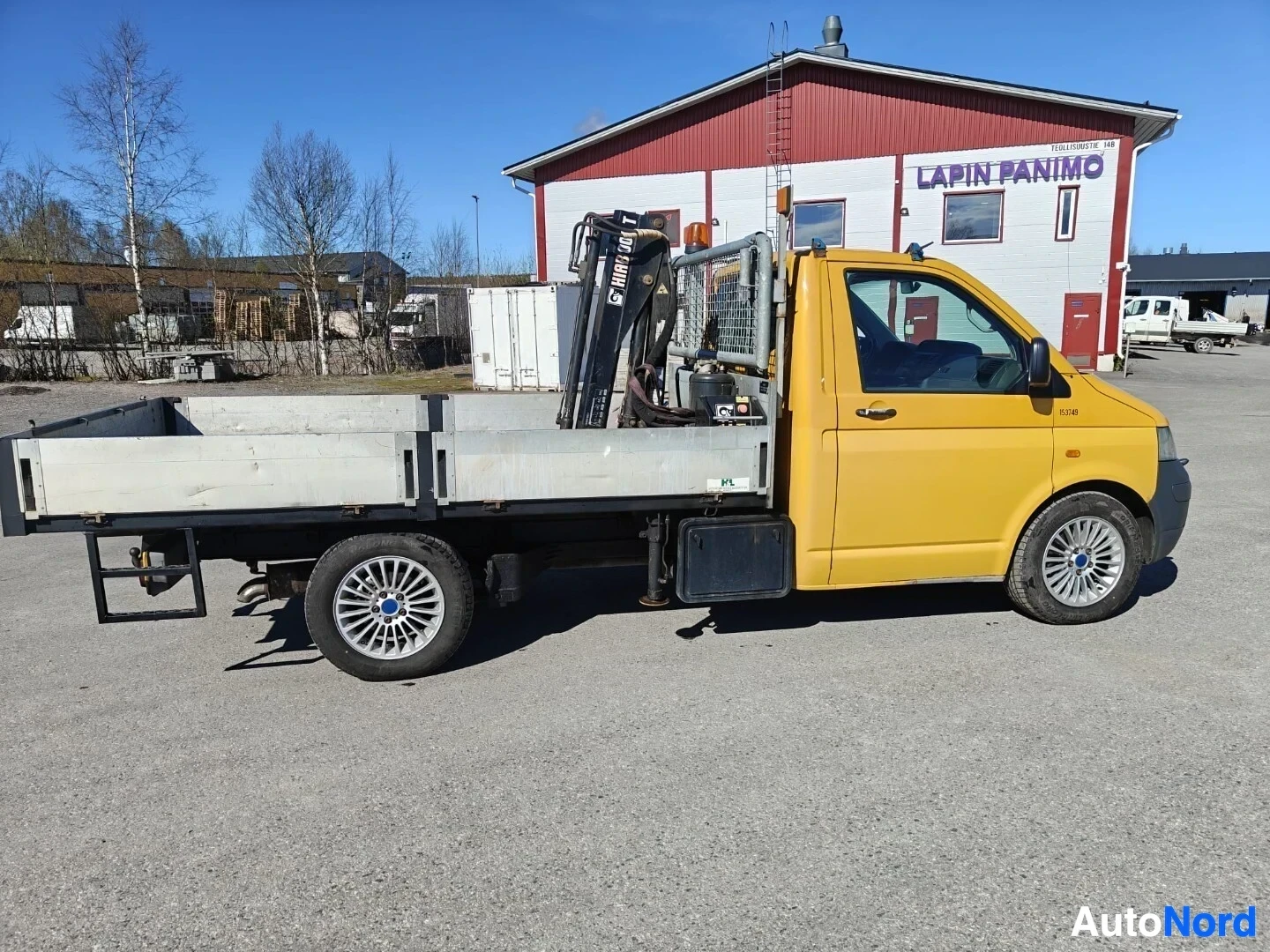 Sivulta: Keltainen avolava-auto, jossa on oranssi varoitusvalo katolla ja musta nosturi lavalla. Ajoneuvossa on kevytmetallivanteet. Taustalla on punainen rakennus, joss