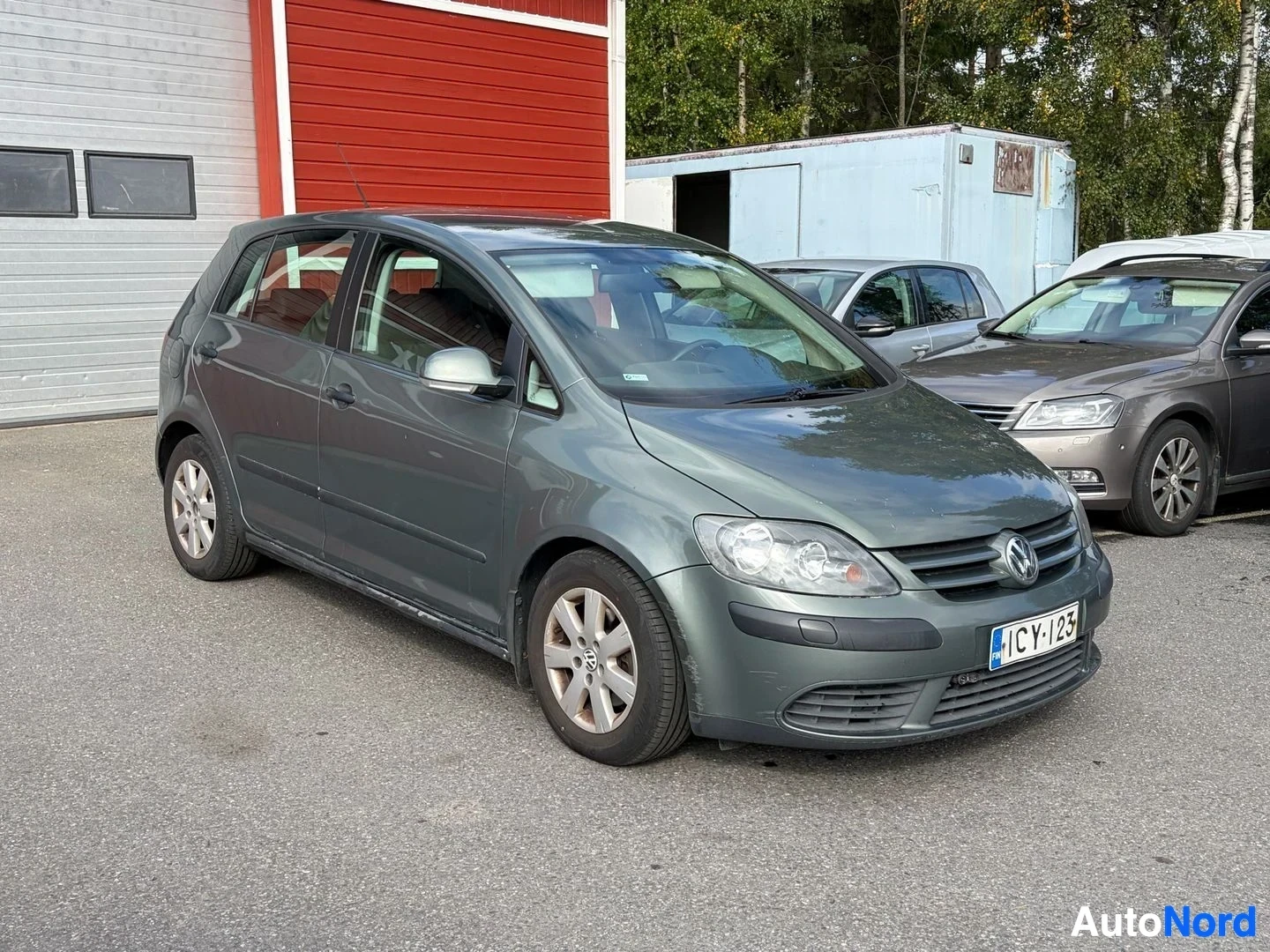 Etuviisto, nokka oikealle: Harmaa Volkswagen Golf Plus -henkilöauto pysäköitynä punaisen rakennuksen ja puuston eteen, rekisterikilpi ""
