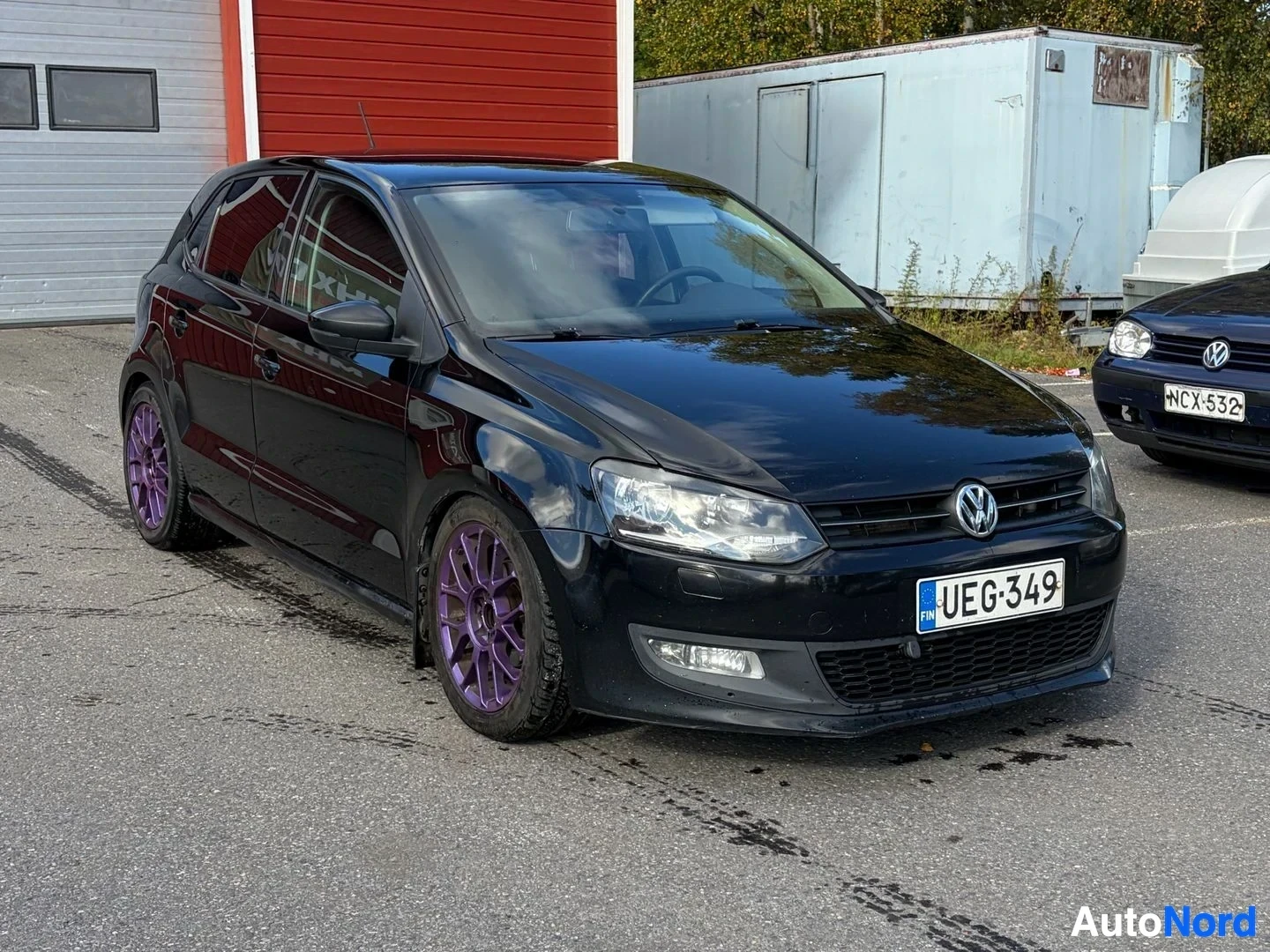 Etuviisto, nokka vasemmalle: Musta auto parkkipaikalla, punainen rakennus takana. Autossa on musta koripaneeli, tummennetut ikkunat, etupuskuri sumuvaloilla ja violetit kevytmetall
