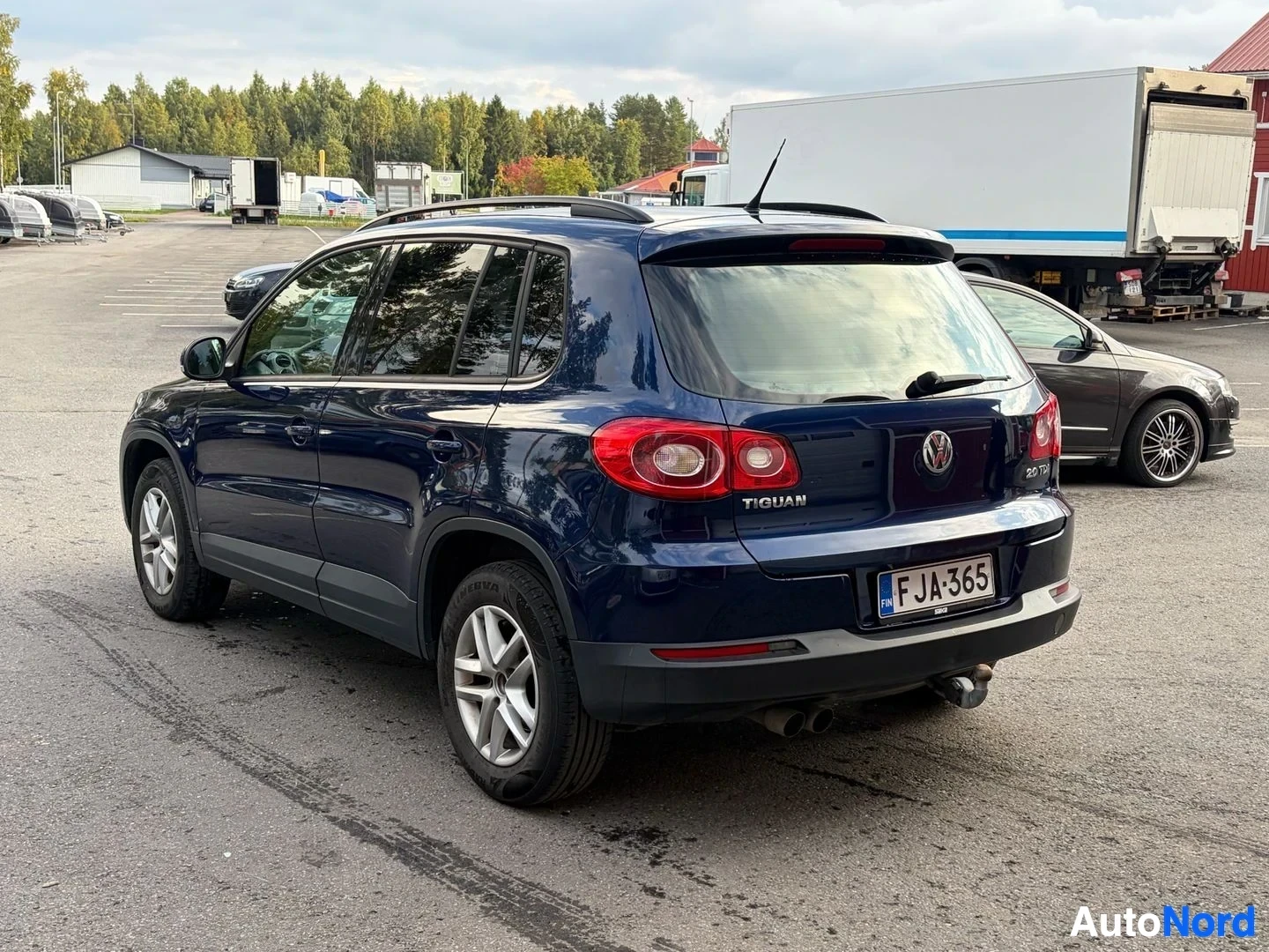 Takaviisto: Sinisessä SUV-ajoneuvossa on kattokaiteet, takana vetokoukku ja suomalainen rekisterikilpi. Takaluukussa lukee 'TIGUAN' ja '2.0 TDI'