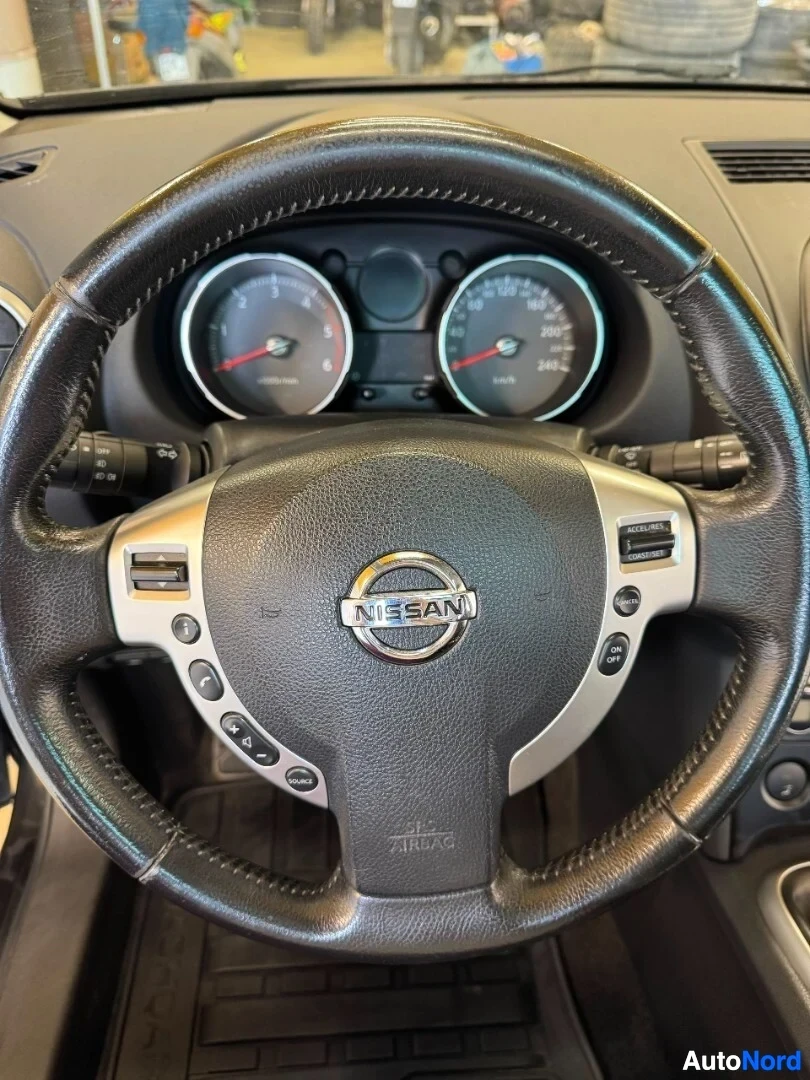Yksityiskohta: Lähikuva auton ohjauspyörästä, jossa on musta nahkaverhoilu, hopeisia yksityiskohtia ja Nissan-logo keskellä. Taustalla näkyy mittaristo.