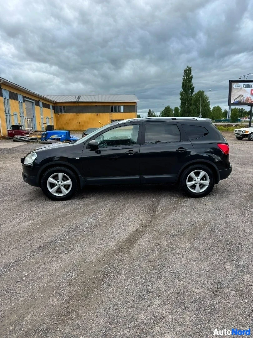 Sivulta: Musta henkilöauto pysäköitynä soraparkkipaikalle rakennuksen eteen, pilvisellä säällä. Auton kyljessä on kattokaiteet ja alumiinivanteet.
