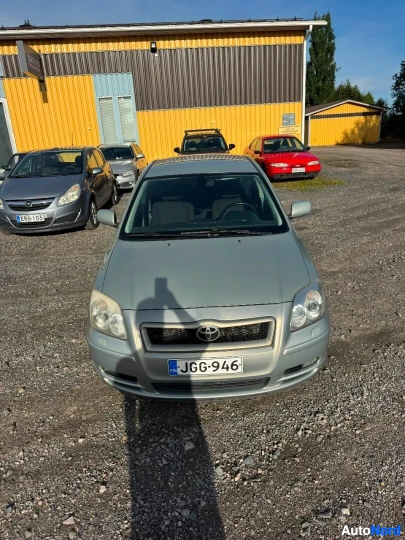 Edestä: Vaaleanharmaa henkilöauto edestä, taustalla kelta-ruskea rakennus ja muita autoja. Ajoneuvon rekisterikilvessä lukee