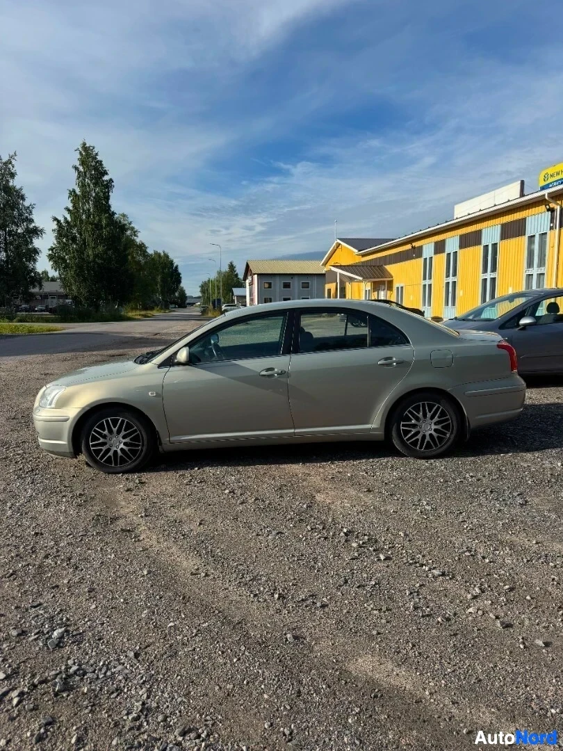Sivulta: Vaaleanharmaa henkilöauto sivusta soraparkkipaikalla. Taustalla näkyy rakennuksia ja puita.