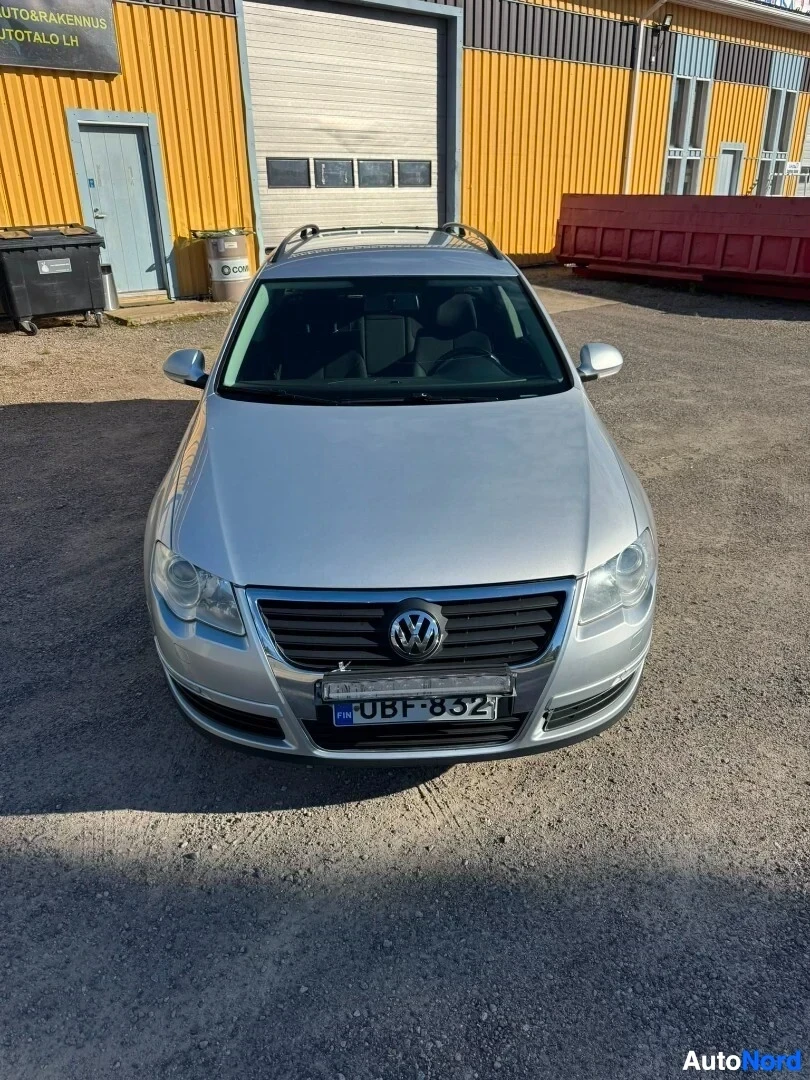 Edestä: Hopeanvärinen farmariauto pysäköitynä sorapihalle rakennuksen eteen. Auto on kuvattu edestäpäin, ja eturekisterikilpi on näkyvissä.