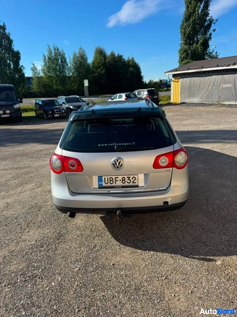 Takaa: Hopeinen Volkswagen Passat Variant -farmariauto kuvattuna suoraan takaa soraparkkipaikalla. Ajoneuvossa on kattokaiteet, takalasin yläpuolella teksti 'www.volks