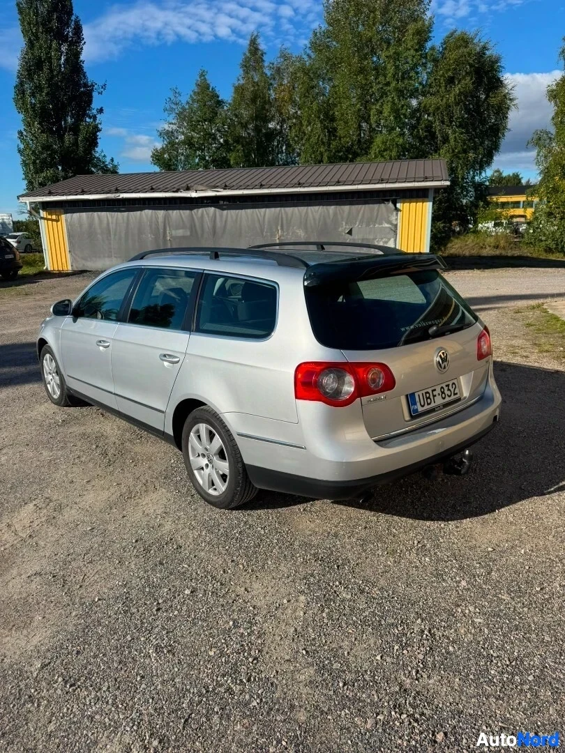 Takaviisto: Hopeinen Volkswagen Passat Variant -farmariauto takaviistosta vasemmalta kuvattuna, pysäköitynä soraparkkipaikalle. Ajoneuvossa on kattokaiteet ja vetokoukku. R