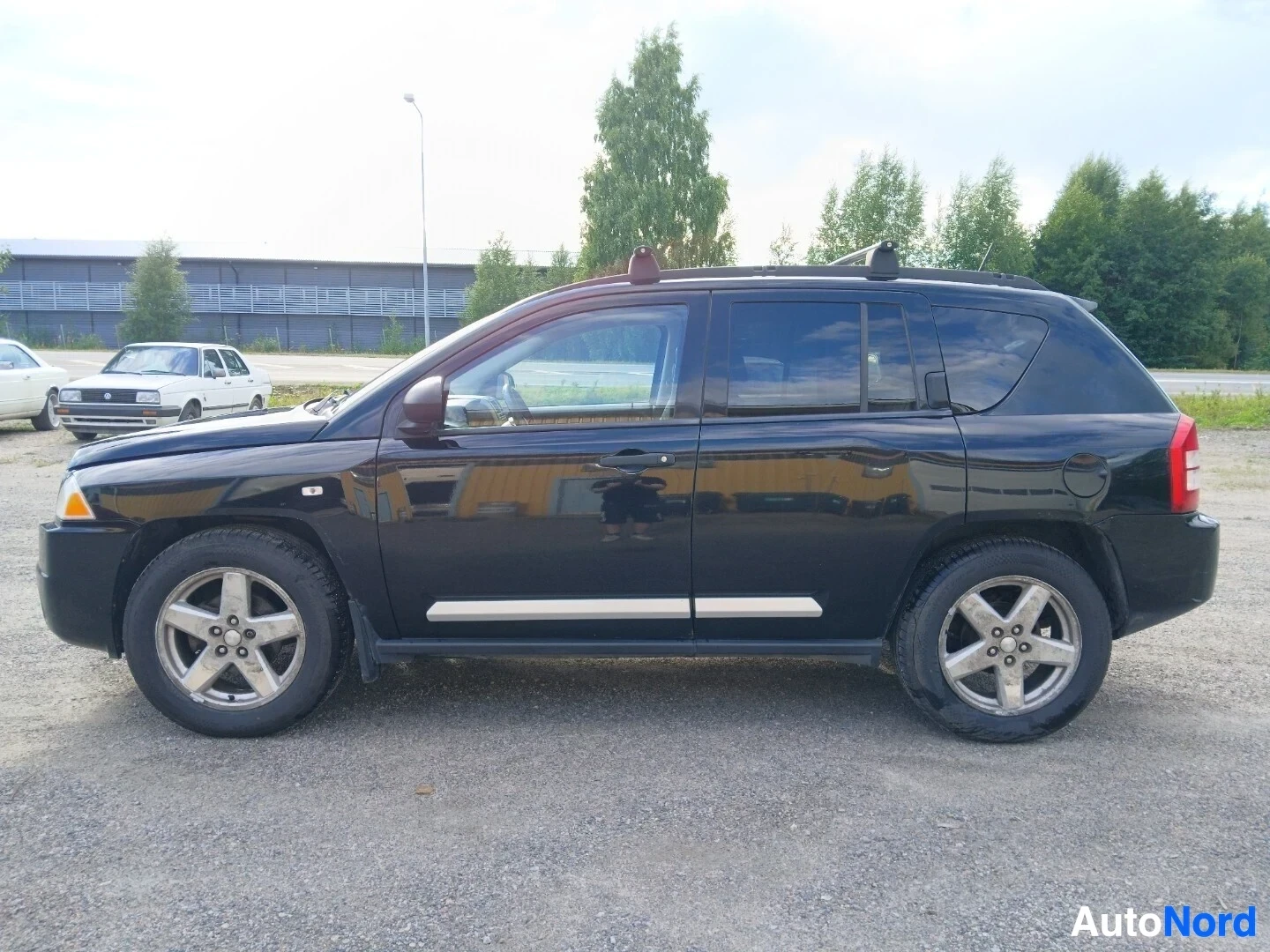 Sivulta: Musta Jeep Compass on kuvattu oikealta puolelta. Autossa on kattotelineet ja viisipuolaiset kevytmetallivanteet.