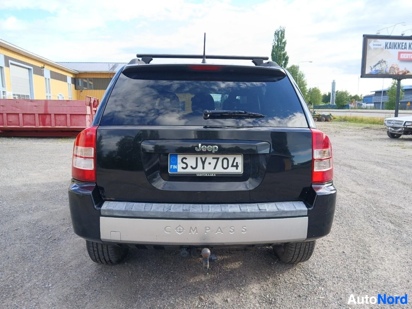 Takaa: Musta Jeep Compass näkyy takaa päin pysäköitynä sorapihalle. Autossa on kattotelineet ja vetokoukku. Rekisterikilpi on luettavissa. Puskurissa on "COMPA