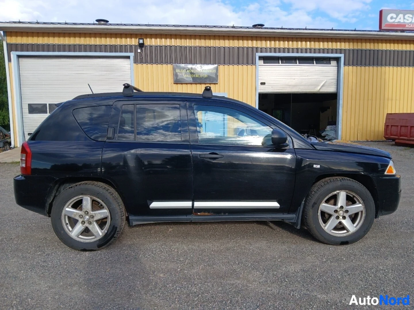 Sivulta: Musta Jeep Compass näkyy vasemmalta sivulta sorapihalla, kaksi ajoneuvon ovea on näkyvissä. Autossa on kattotelineet ja kevytmetallivanteet.
