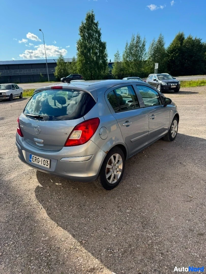 Takaviisto: Harmaa Opel Corsa on pysäköity sorapihalle rakennuksen eteen, takaosasta kuvattuna. Takaosa ja vasen kylki ovat näkyvissä. Rekisterikilpi "" on auton tak