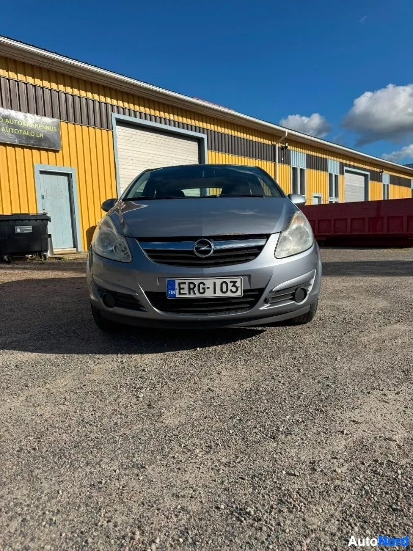 Edestä: Harmaa henkilöauto pysäköitynä sorapihalle kelta-harmaata rakennusta vasten. Auton etuosa on kuvassa, sisältäen etusäleikön, ajovalot ja etupuskurin, sekä rekis