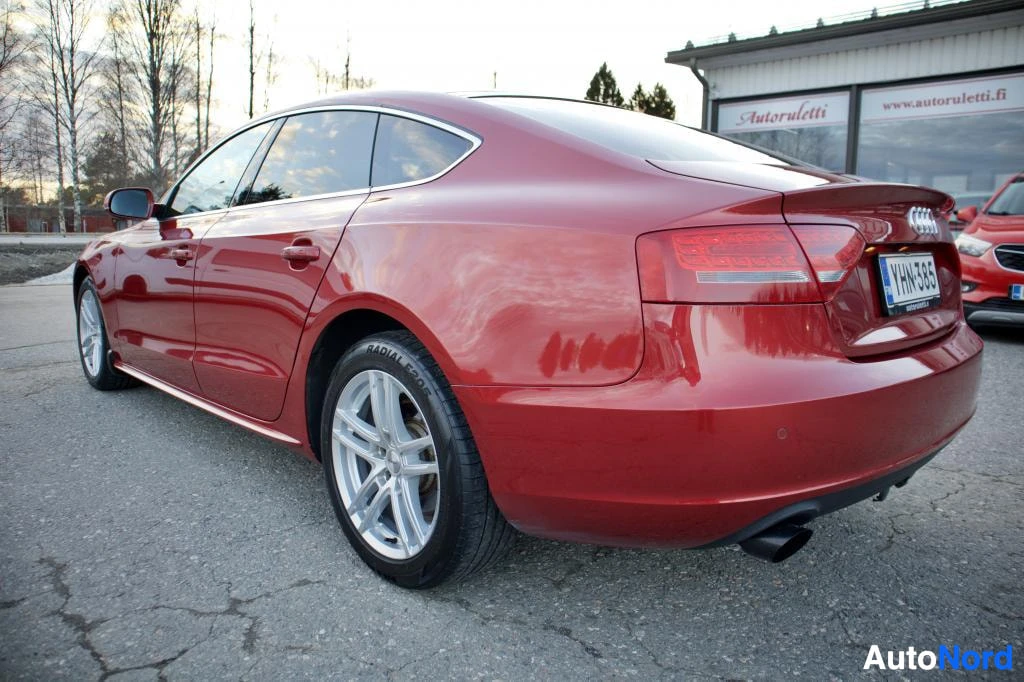 Takaviisto: Punaisen Audi A5 Sportbackin takaosa ja oikea kylki ovat kuvassa. Autossa on hopeiset kevytmetallivanteet. Rekisterikilpi on osittain näkyvissä. Taustalla näkyy