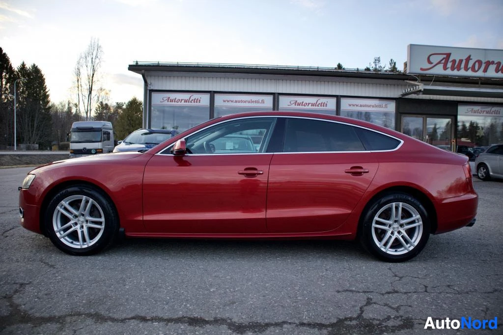 Sivulta: Punainen Audi A5 Sportback on pysäköitynä tasaisella alustalla, kuvattuna oikealta sivulta. Autossa on hopeiset monipuolaiset kevytmetallivanteet. Taustalla näk