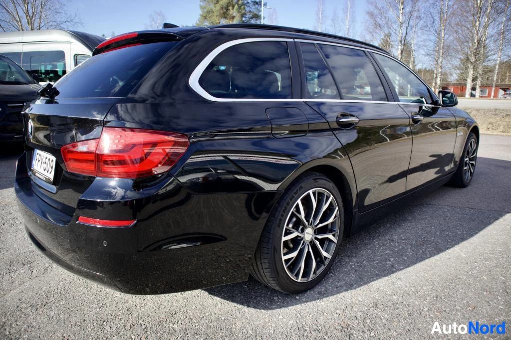 Takaviisto: Musta BMW 5-sarjan farmari kuvattuna takavasemmalta viistosti. Takarekisterikilpi "" ja takavalot ovat näkyvissä. Autossa on kattokaiteet ja monipuolaiset kevyt