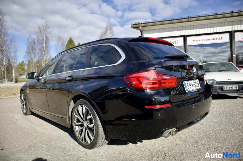 Takaviisto: Musta BMW 5-sarjan farmari kuvattuna takaoikealta viistosti. Rekisterikilpi "", takavalot ja kattokaiteet ovat näkyvissä. Autossa on monipuolaiset kevytmetalliv