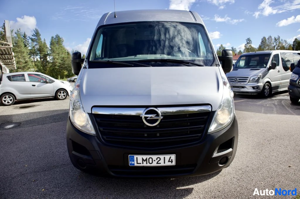 Edestä: Hopeanvärinen pakettiauto, jonka keula on kuvassa. Ajoneuvossa on suomalainen rekisterikilpi. Taustalla näkyy muita ajoneuvoja ja puita.