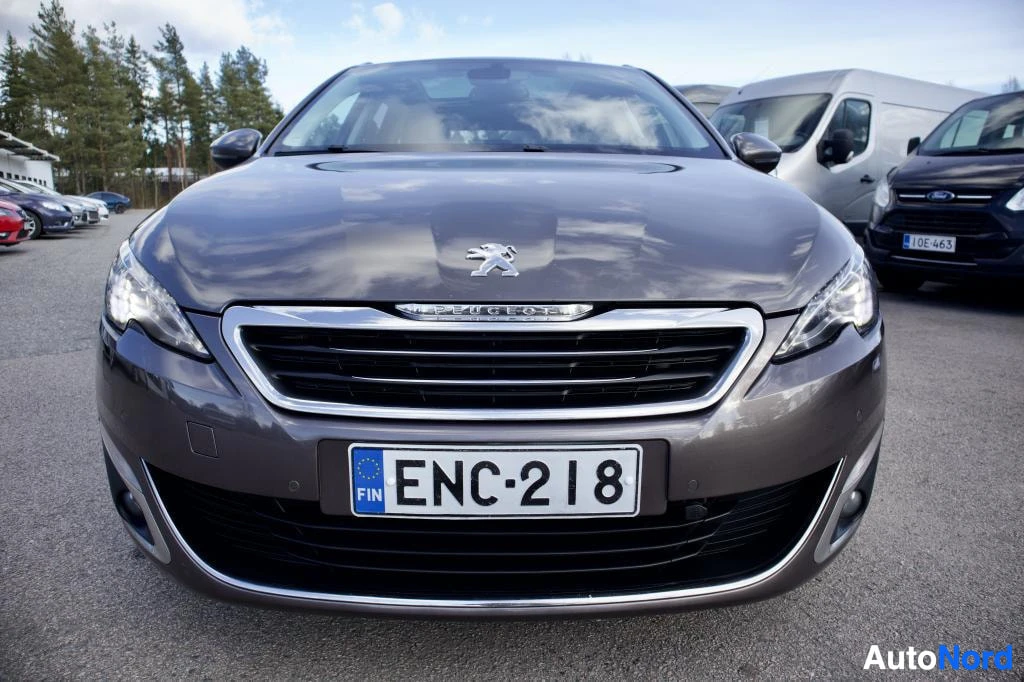 Edestä: Auton etuosa edestäpäin kuvattuna. Tummanruskea Peugeot-merkkinen auto on pysäköitynä asfaltille. Sen etusäleikkö on kromattu ja puskurin alaosassa on musta rit