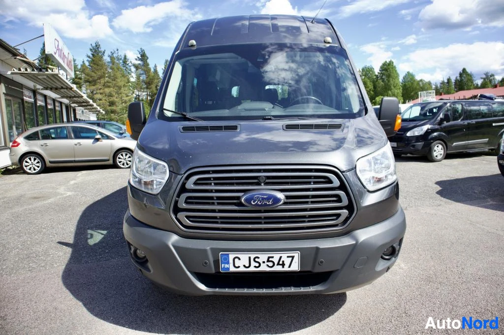 Edestä: Tummanharmaa pakettiauto pysäköitynä ulos, edestä kuvattuna. Rekisterikilpi näkyy. Auton etusäleikkö, ajovalot ja etupuskuri ovat kuvassa. Taustalla on muita aj
