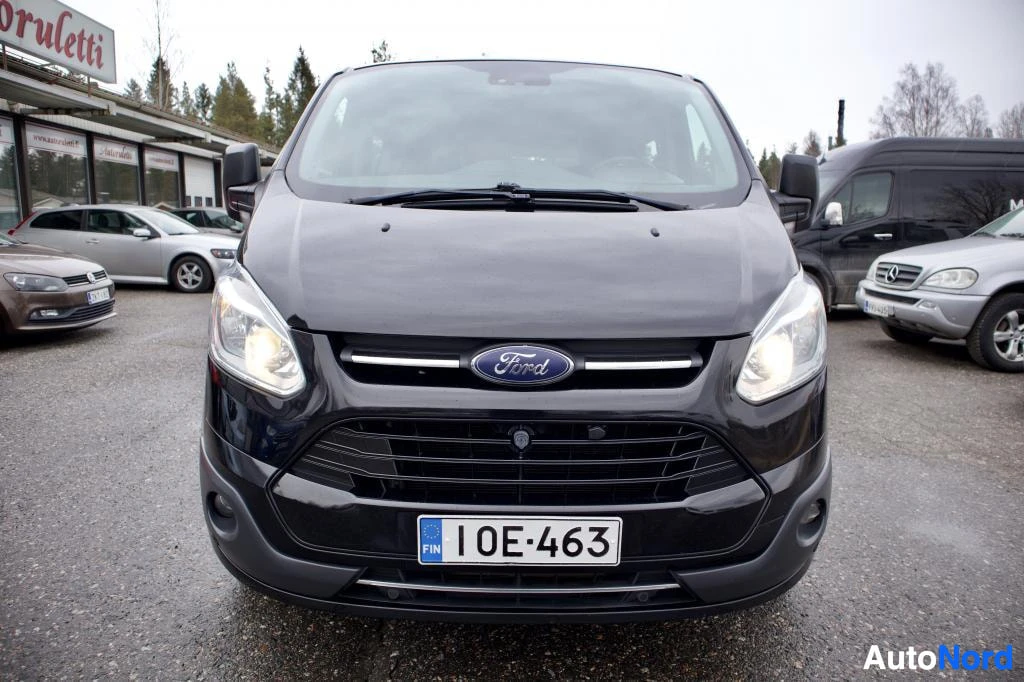 Edestä: Musta pakettiauto edestäpäin kuvattuna, ajovalot päällä.