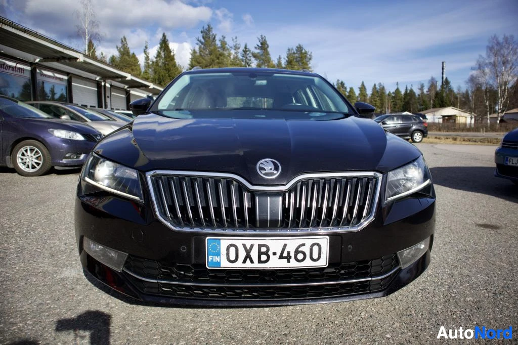 Edestä: Mustan Skoda Superb -farmariauton etuosa, jossa näkyvät säleikkö, ajovalot ja etupuskuri. Rekisterikilpi "" on keskitettynä