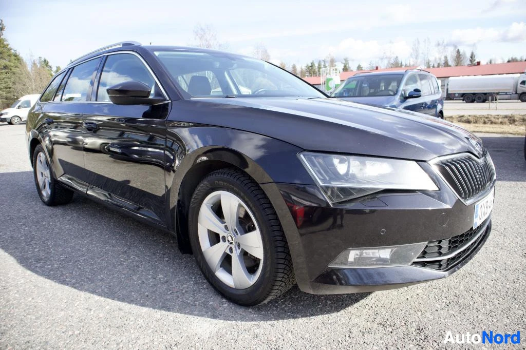Etuviisto, nokka oikealle: Mustan Skoda Superb -farmariauton etuvasen puoli, jossa näkyvät ajovalot, kevytmetallivanne ja ikkunat. Auto on pysäköity asfaltoidulle alueelle.