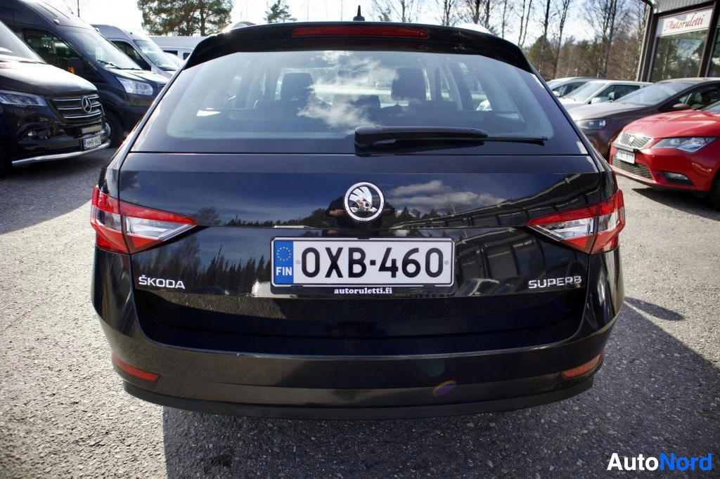 Takaa: Auton takaosa, jossa on rekisterikilpi "", Skoda-logo ja Superb-teksti. Tumman sininen maalipinta heijastaa pilvisiä taivaita