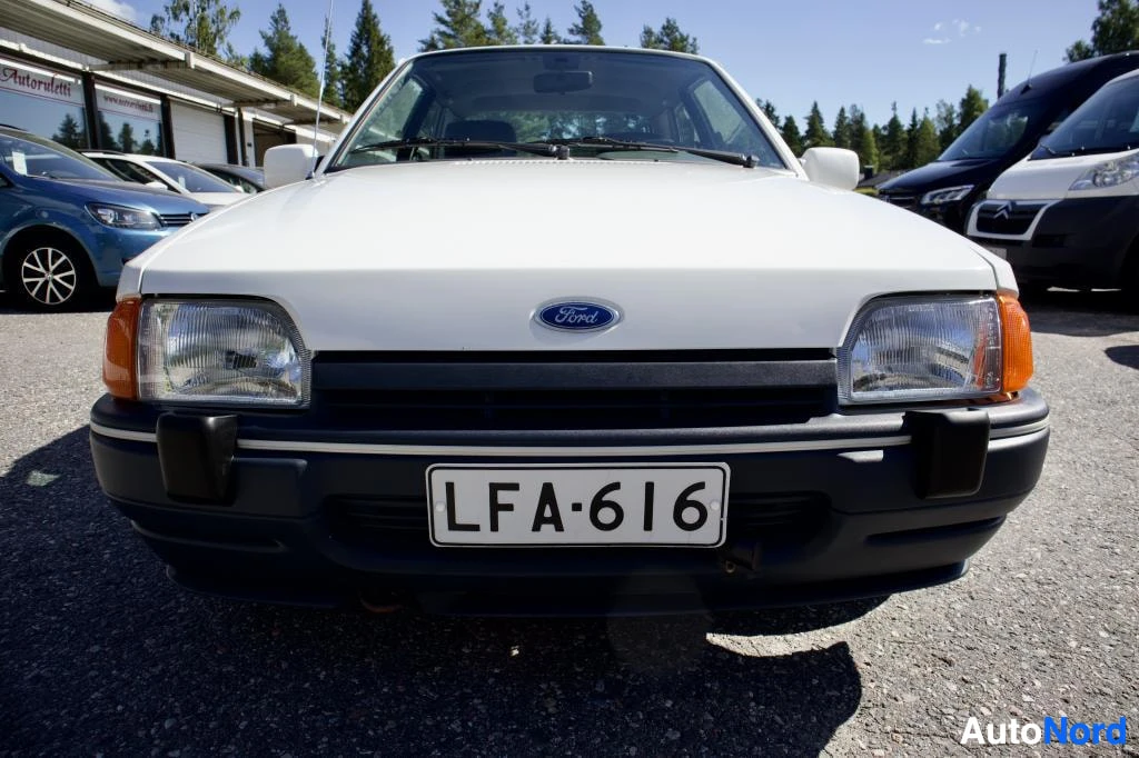 Edestä: Valkoisen Ford Escortin keula pysäköintialueella. Autossa on valkoinen rekisterikilpi "" ja sininen Ford-logo jäähdyttimen säleikön yläpuolella. Ajoneuvossa on