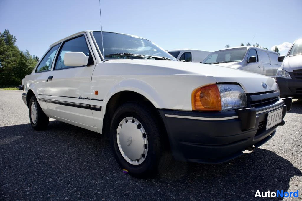 Etuviisto, nokka oikealle: Valkoinen Ford Escort pysäköitynä ulkona aurinkoisella säällä. Auton keula ja oikea kylki ovat näkyvissä. Autossa on tummanharmaat puskurit ja kylkilis