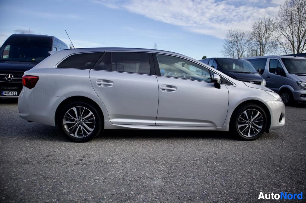 Sivulta: Hopeanvärinen farmariauto seisoo ulkona vasemmalta sivulta kuvattuna. Takana näkyy myös musta pakettiauto, jonka rekisterikilpi on ""