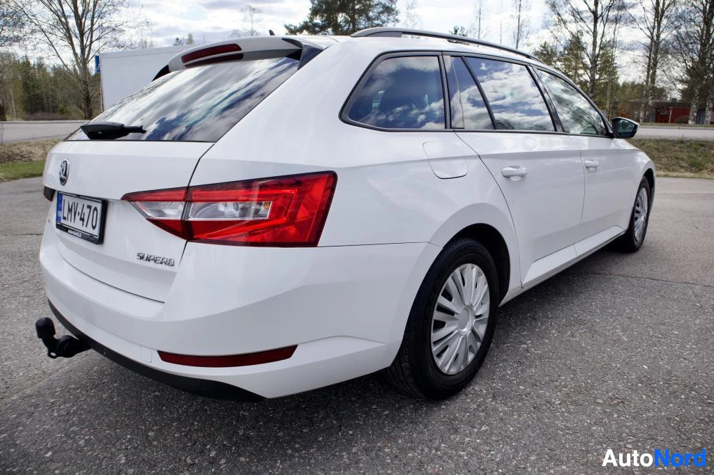 Takaviisto: Valkoinen Skoda Superb Combi, jossa on vetokoukku ja kattokiskot, pysäköitynä. Ajoneuvon vasen takakulma on näkyvissä.