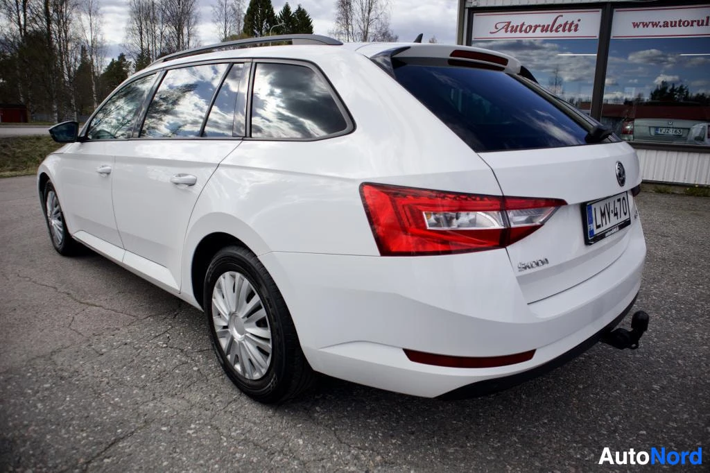 Takaviisto: Valkoinen Skoda Superb Combi -farmariauto, jossa on vetokoukku ja kattokiskot, pysäköitynä. Ajoneuvon oikea takakulma on näkyvissä.