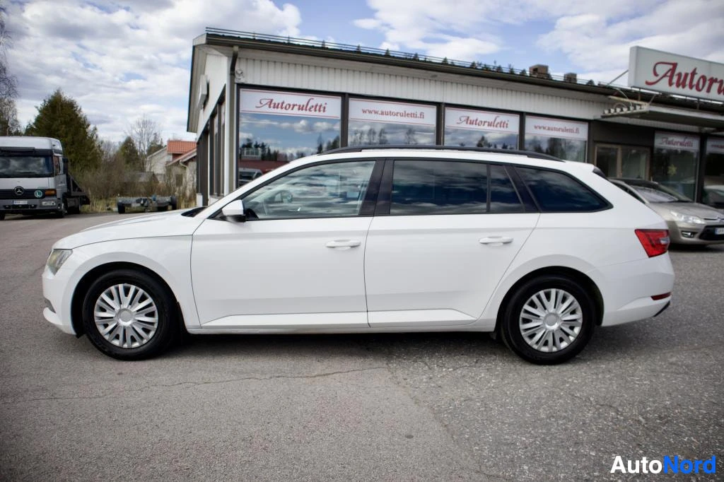 Sivulta: Valkoinen Skoda Superb Combi -farmariauto, jossa on kattokiskot ja kevytmetallivanteet, pysäköitynä tasaisella alustalla. Ajoneuvo on kuvattu oikealta sivulta.