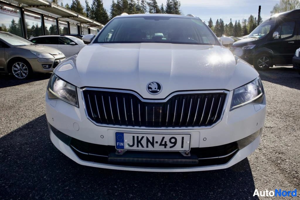 Edestä: Valkoisen farmariauton etuosa on kuvattu suoraan edestä. Rekisterikilpi on näkyvissä. Etuvalot ovat päällä. Auto on pysäköity ulos, ja taustalla näkyy m