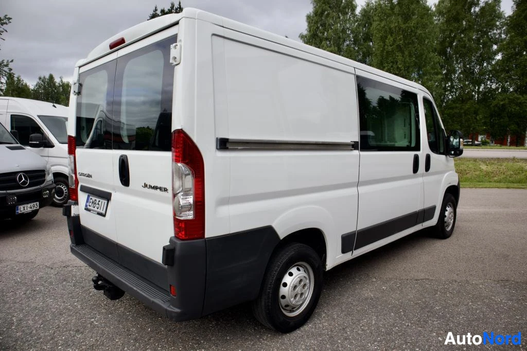 Takaviisto: Valkoinen pakettiauto, jossa on takaluukussa kaksi ikkunaa, perävaunun vetokoukku ja teräsvanteet, on kuvattu takaviistosta. Auton rekisterikilpi on.