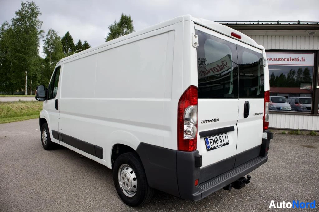 Takaviisto: Valkoinen pakettiauto, jossa on takaluukussa kaksi ikkunaa, perävaunun vetokoukku ja teräsvanteet, on kuvattu takaviistosta. Rekisterikilpi on.