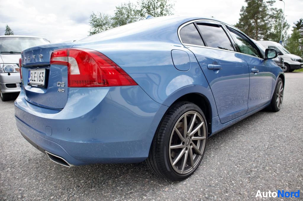 Takaviisto: Sininen Volvo S60 -henkilöauto pysäköitynä ulkona, kuvattuna takaviistosta oikealta puolelta. Autossa on hopeanväriset monipuolaiset kevytmetallivanteet ja taka