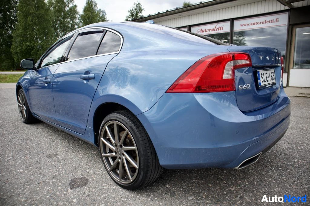 Takaviisto: Sininen Volvo S60 -henkilöauto pysäköitynä ulkona, kuvattuna takaviistosta vasemmalta puolelta. Autossa on hopeanväriset monipuolaiset kevytmetallivanteet ja ta