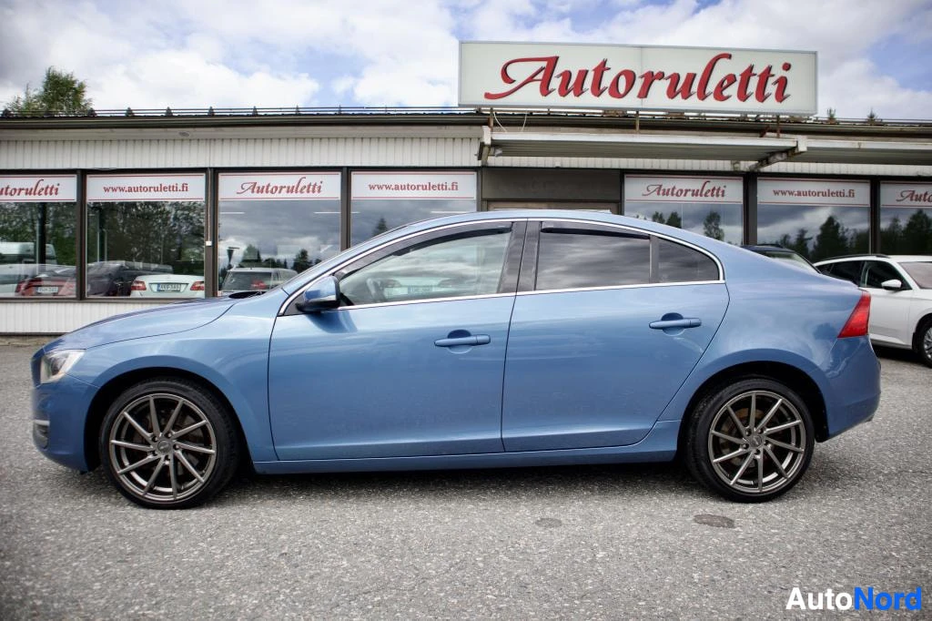 Sivulta: Sininen Volvo S60 -henkilöauto pysäköitynä ulkona, kuvattuna vasemmalta sivulta. Autossa on hopeanväriset monipuolaiset kevytmetallivanteet ja tummennetut sivui