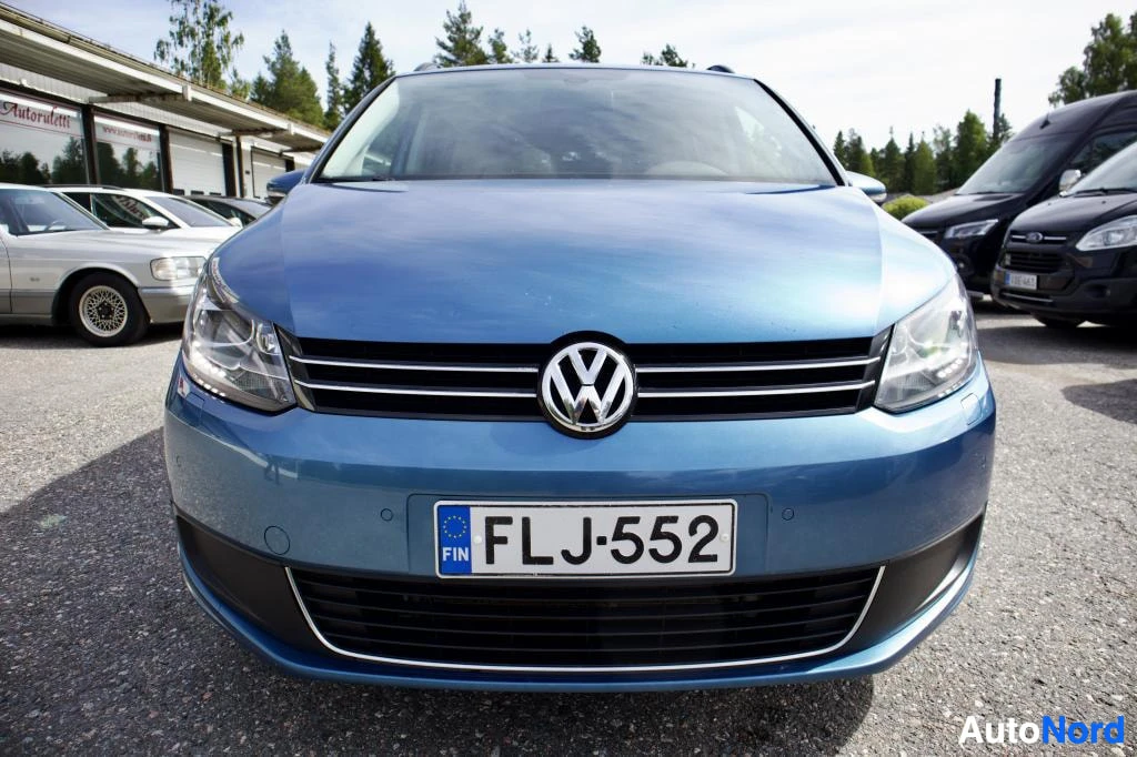 Edestä: Sinisessä VW Touranissa on rekisterikilpi. Auton keula on kuvassa keskellä ja etupuskurissa näkyy pysäköintitutkan anturit. Etusäleikkö on krominen ja s