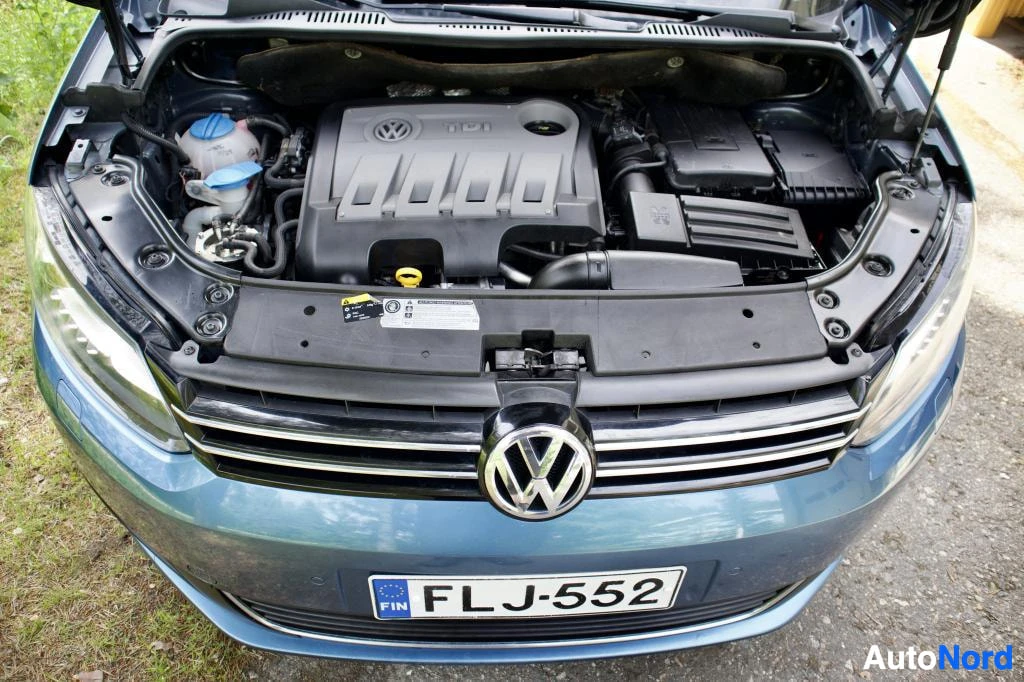Moottoritila: Ajoneuvon konepelti on auki ja moottori on näkyvissä. Keulan alaosa, jossa on rekisterikilpi ja Volkswagen-logo, on myös kuvassa. Rekisterikilven teksti on FLJ-