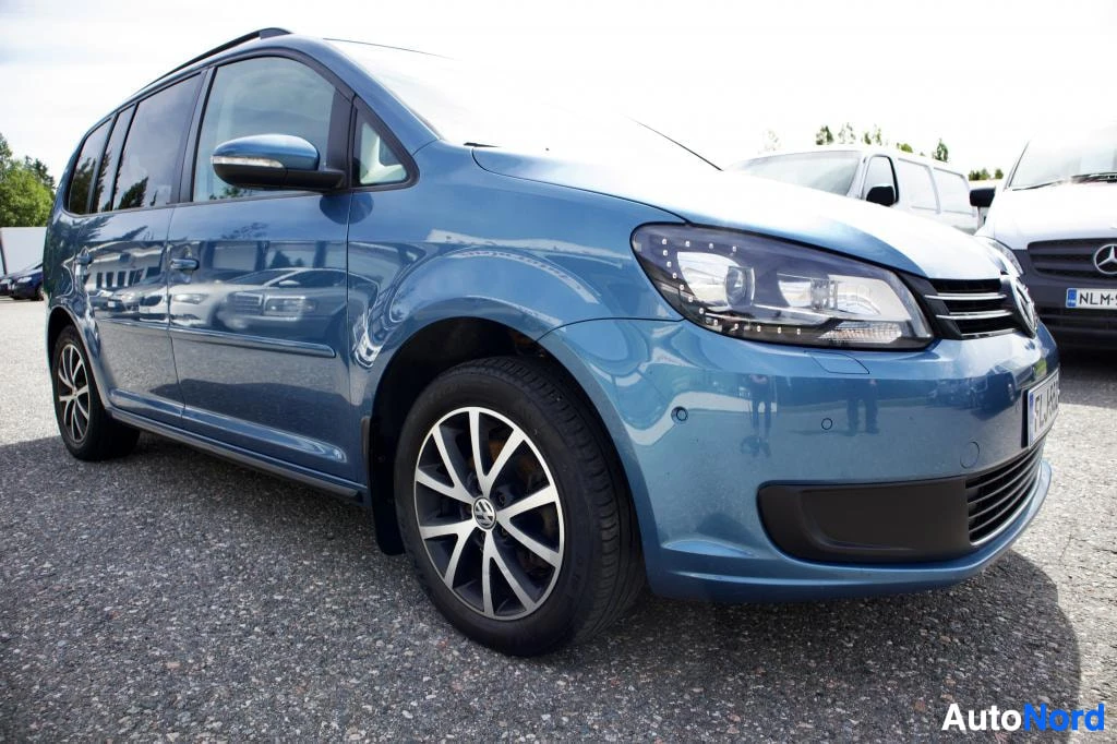 Etuviisto, nokka oikealle: Sinisessä VW Touranissa on tummat ikkunat. Auton keula on kuvassa oikealla ja vasen sivupaneeli näkyy. Edessä rekisterikilvessä teksti. Auton ala