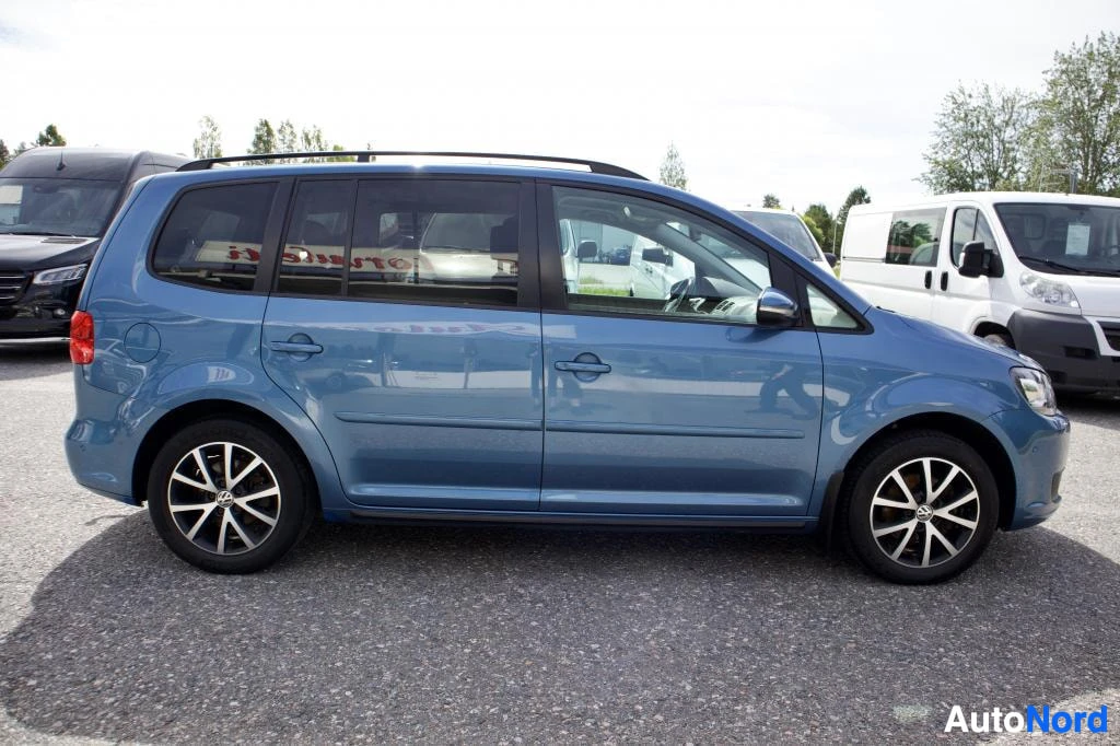 Sivulta: Sinisessä VW Touranissa on tummat ikkunat ja kattokaiteet. Auton vasen kylki näkyy kokonaan. Ajoneuvossa on monipuolaiset kevytmetallivanteet. Auto on pysäköity