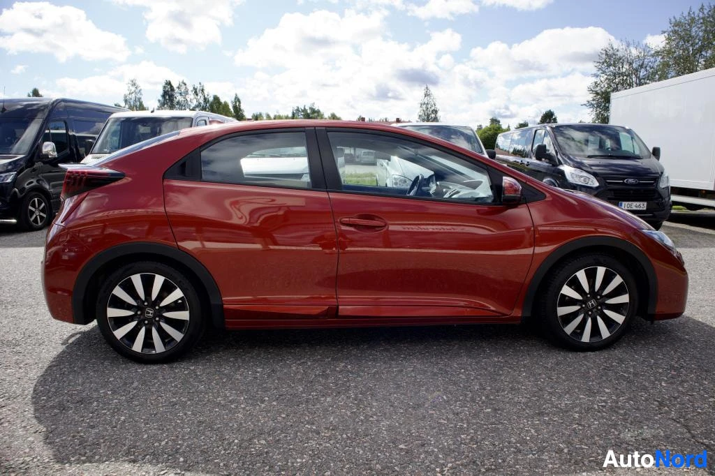 Sivulta: Punainen henkilöauto oikealta sivulta. Auto on pysäköity ulos ja taustalla näkyy muita ajoneuvoja ja rakennus. Autossa on musta-hopeiset kevytmetallivanteet.