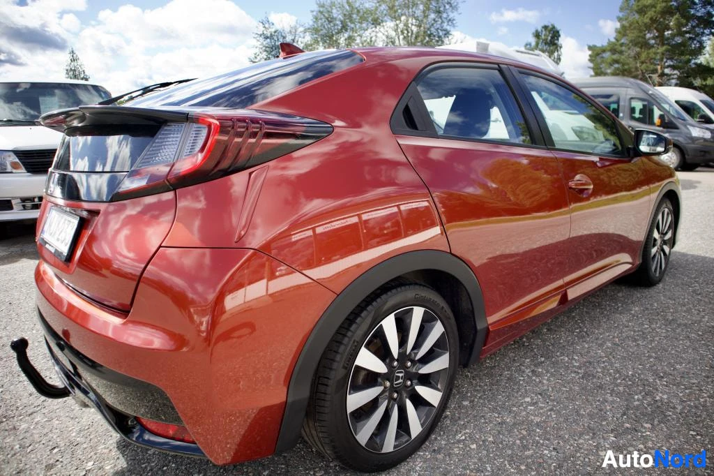 Takaviisto: Oranssinpunainen henkilöauto, jossa on tumma takaluukku ja musta takapuskuri, pysäköitynä ulkona päivänvalossa. Kuvassa näkyy auton takaosa ja kylki, ja autossa